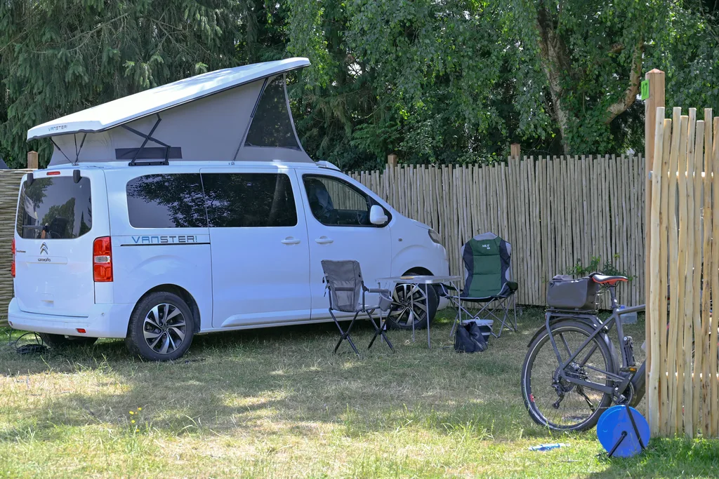 Van aménagé au toit relevable garé sur un emplacement nu, chaises et vélo, palissade en châtaignier